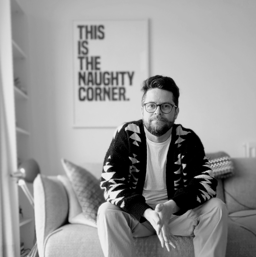 Paul Mackenzie, creative director, seated on a sofa in front of a print that reads 'This is the naughty corner.'